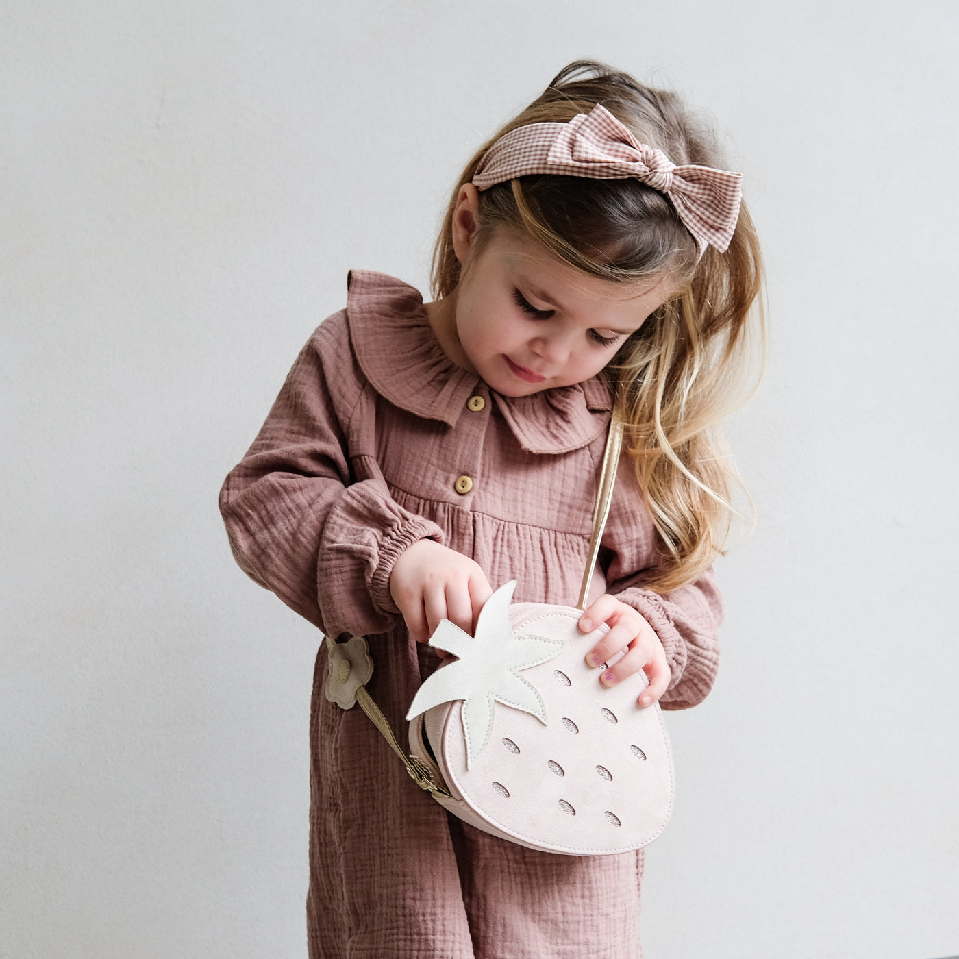 Little girl wearing pretty bow, gingham print brown Alice band opening a strawberry shape bag