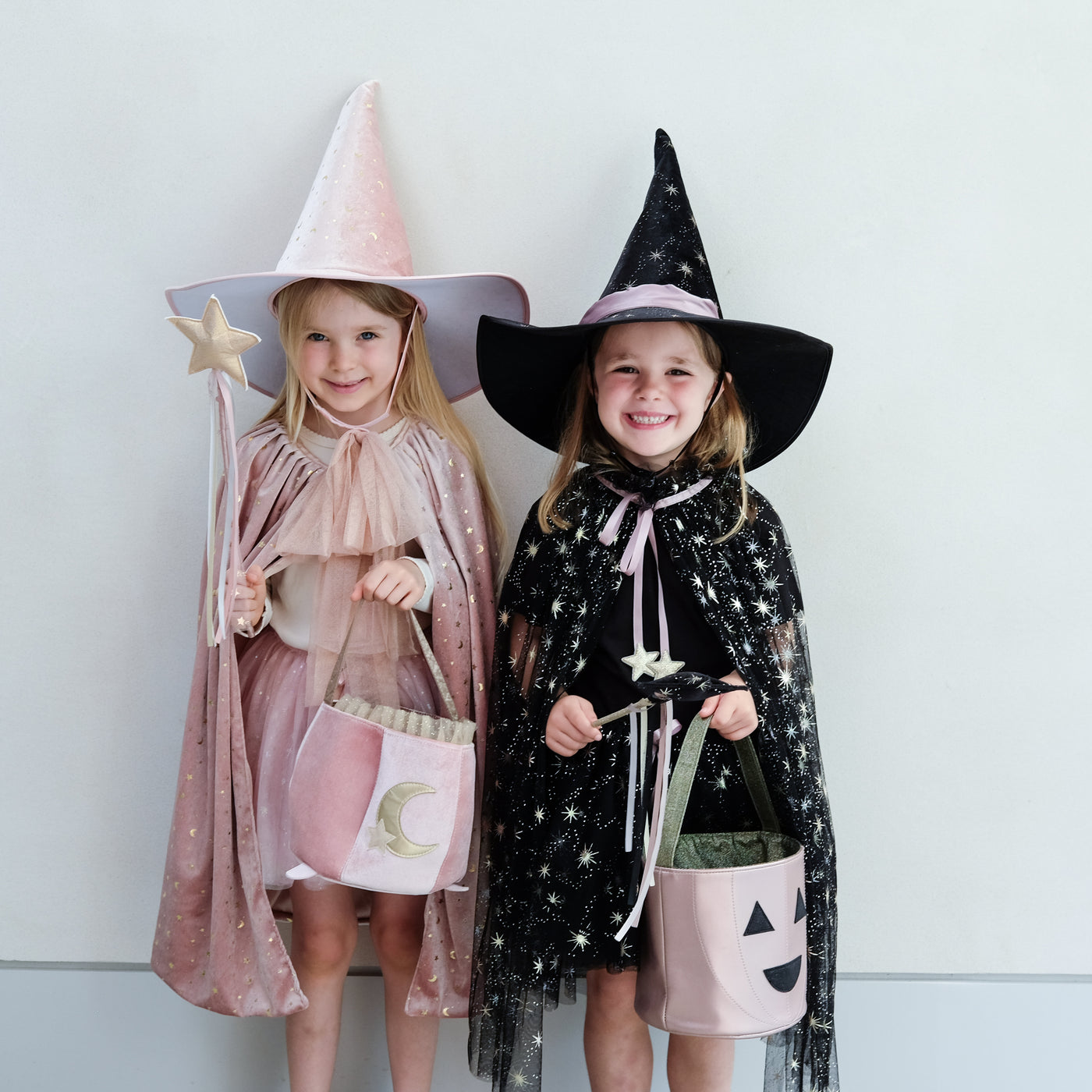 Two little girls in witch costumes, one soft pink velvet, one black tulle and velvet with star print, both holding trick or treat bags in pumpkin and cauldron designs