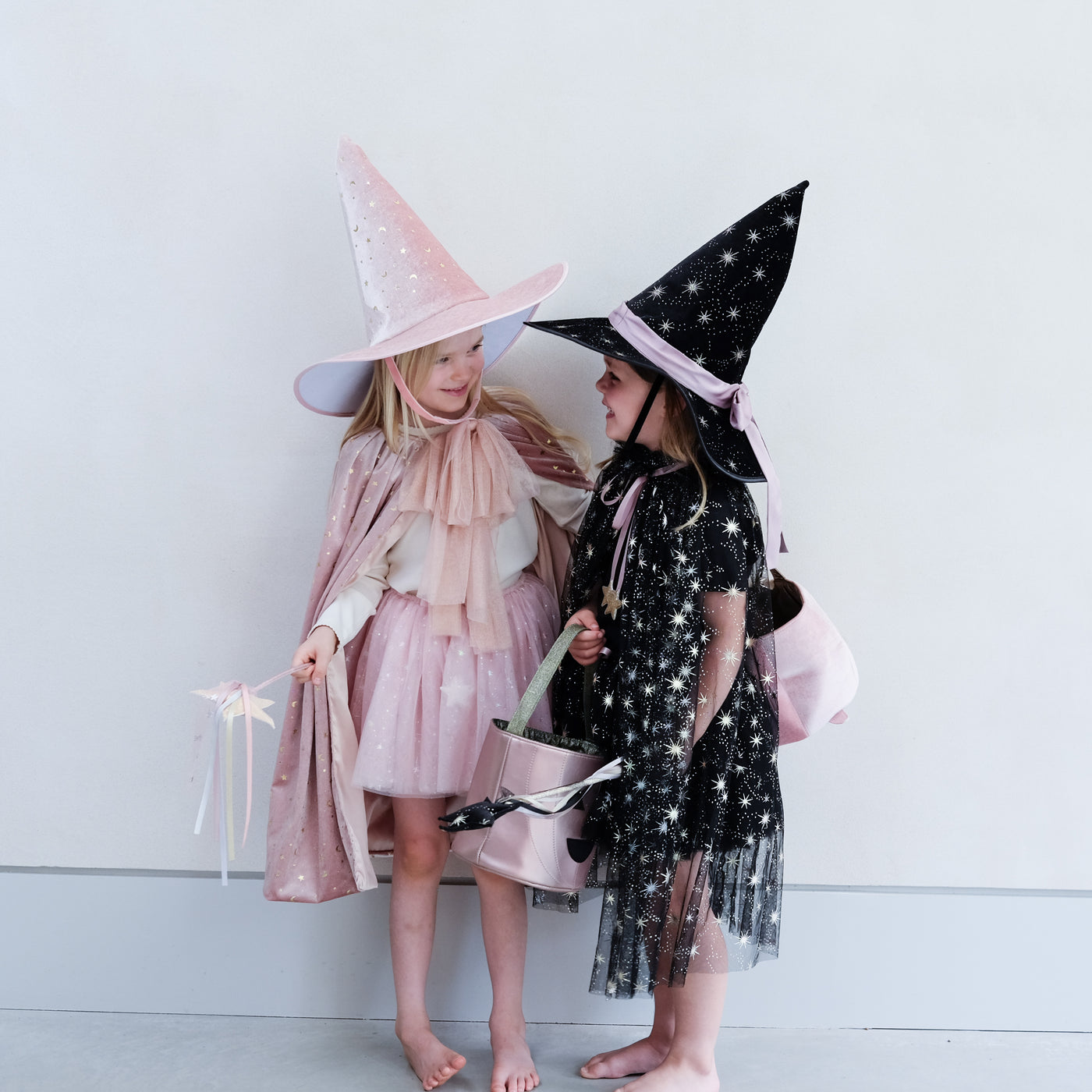 Two little girls in witch costumes, one soft pink velvet, one black tulle and velvet with star print, both holding trick or treat bags in pumpkin and cauldron designs