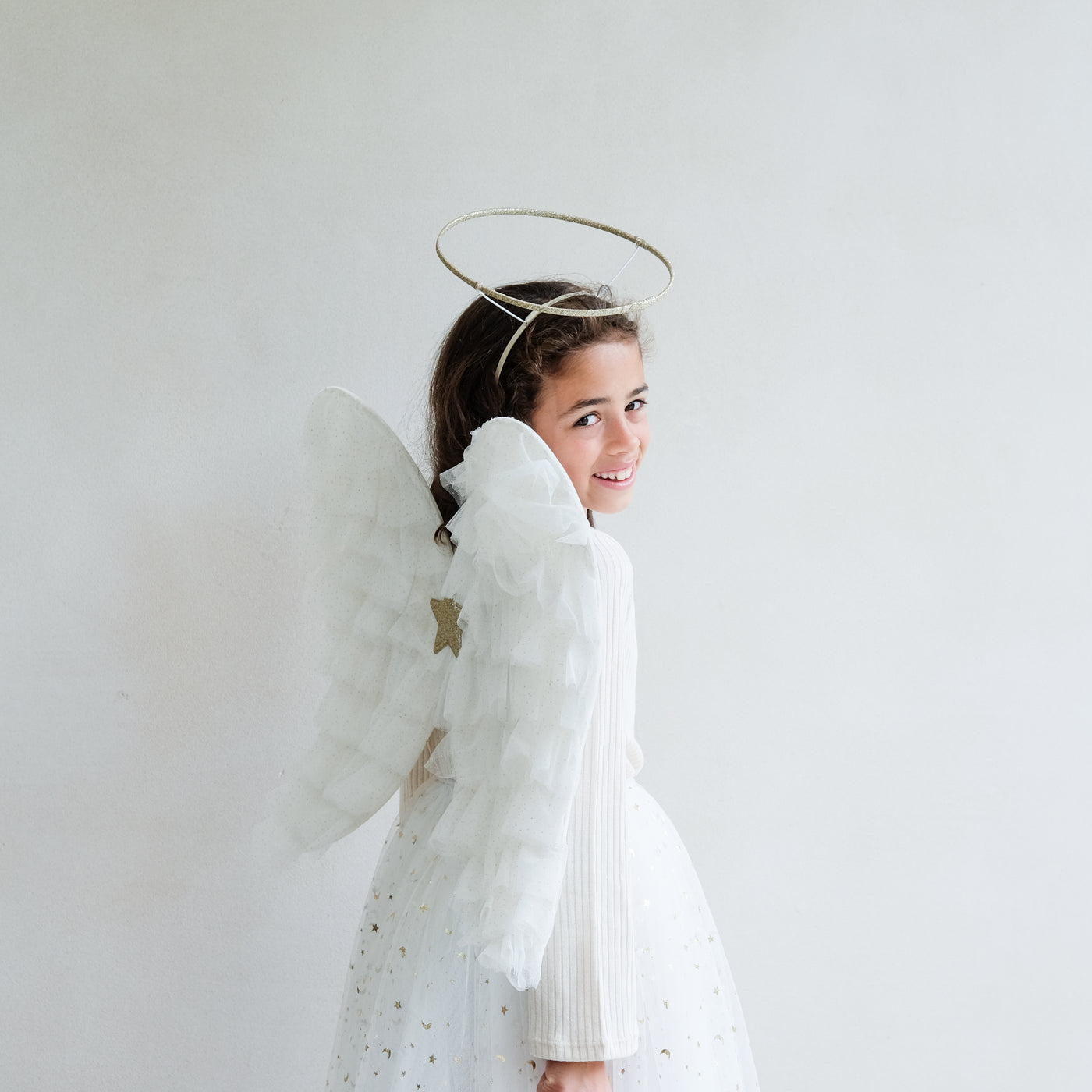 Dark haired little girl wearing a white tulle layered tutu, a gold halo headdress and a beautiful pair of angel wings