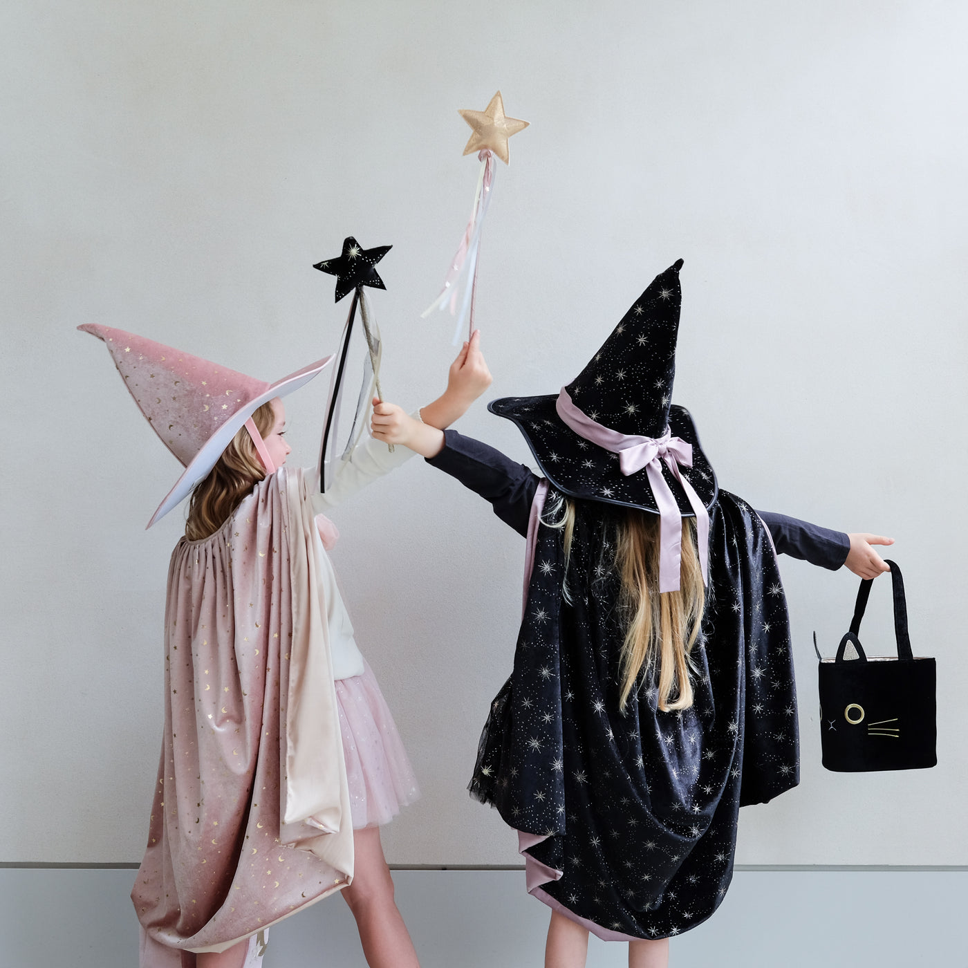Two little girls dressed as witches, one in pink, one in black with star wands with ribbon trails and a black velvet cat trick or treat bag