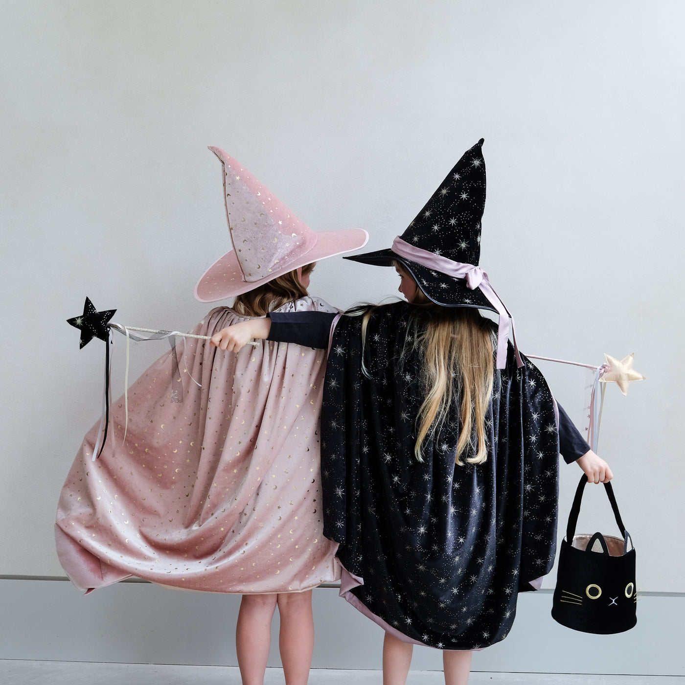 Two little girls dressed as witches, one in pink, one in black with star wands with ribbon trails and a black velvet cat trick or treat bag