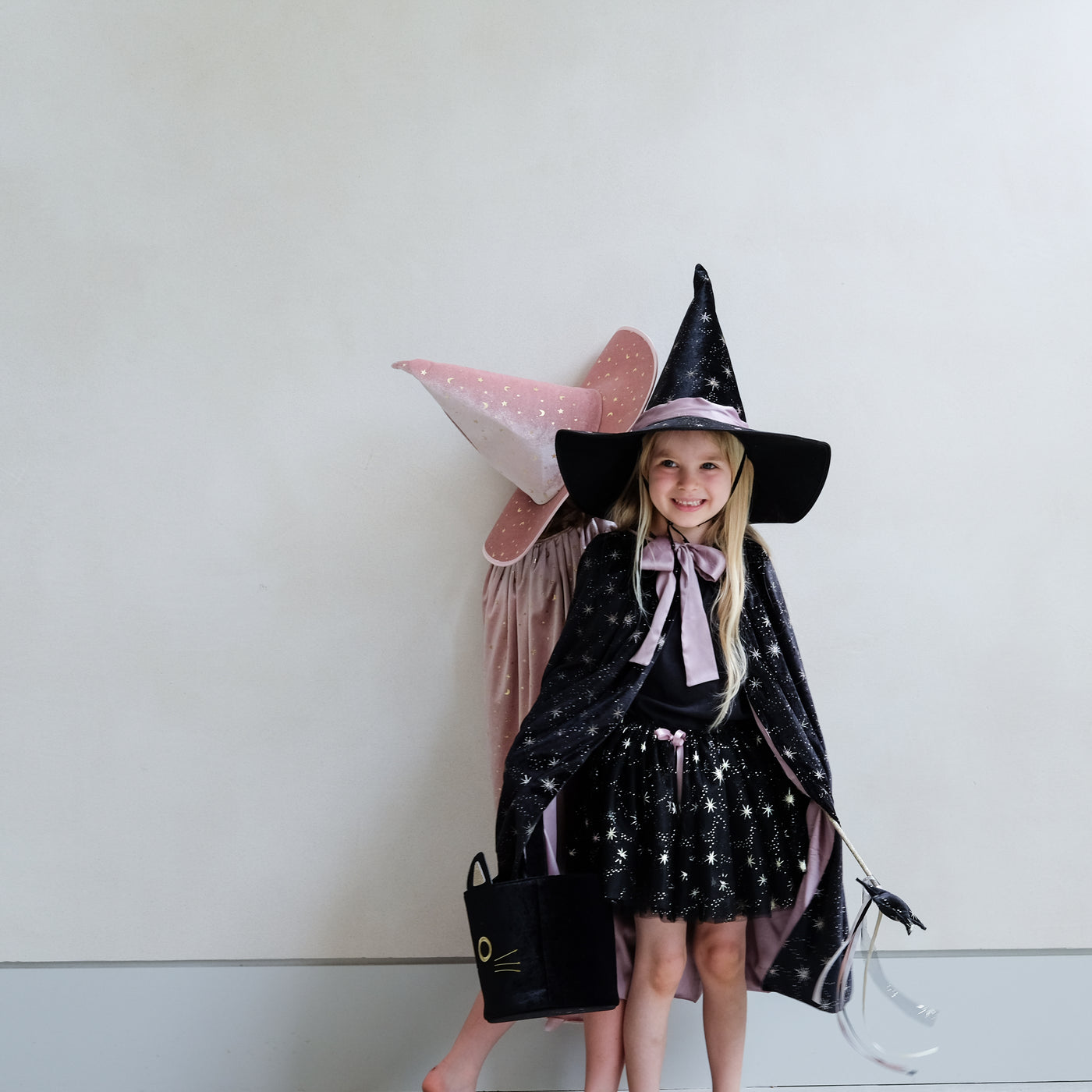 Two little girls dressed as witches, one in pink, one in black with star wands with ribbon trails and a black velvet cat trick or treat bag