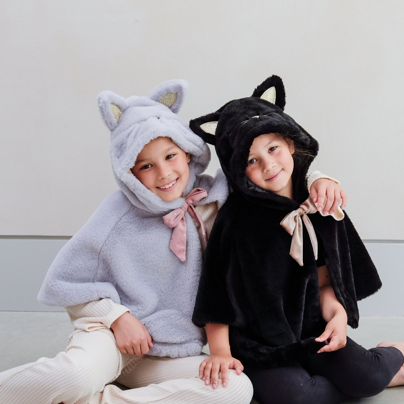 Two children wearing cat capes, one grey and one black, sitting on a light-coloured floor.