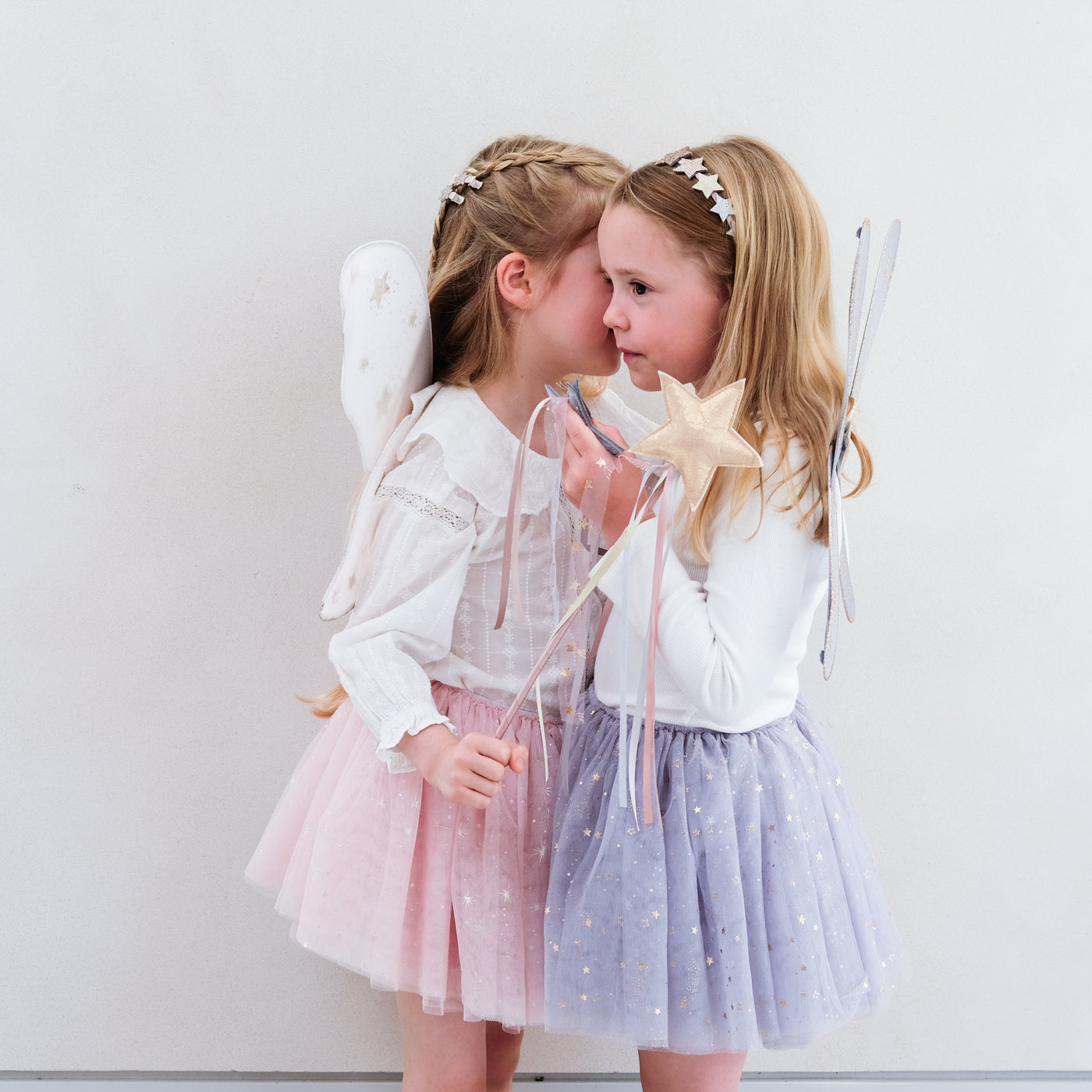 Little girl wearing grey tulle tutu speckled with gold stars and grey netted gold star print fairy wings and another wearing a pink tutu and pink wings, both with matching wands