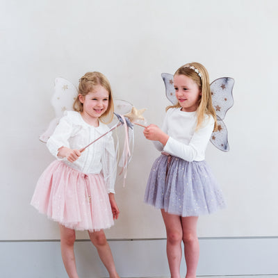 Little girl wearing grey tulle tutu speckled with gold stars and grey netted gold star print fairy wings and another wearing a pink tutu and pink wings, both with matching wands