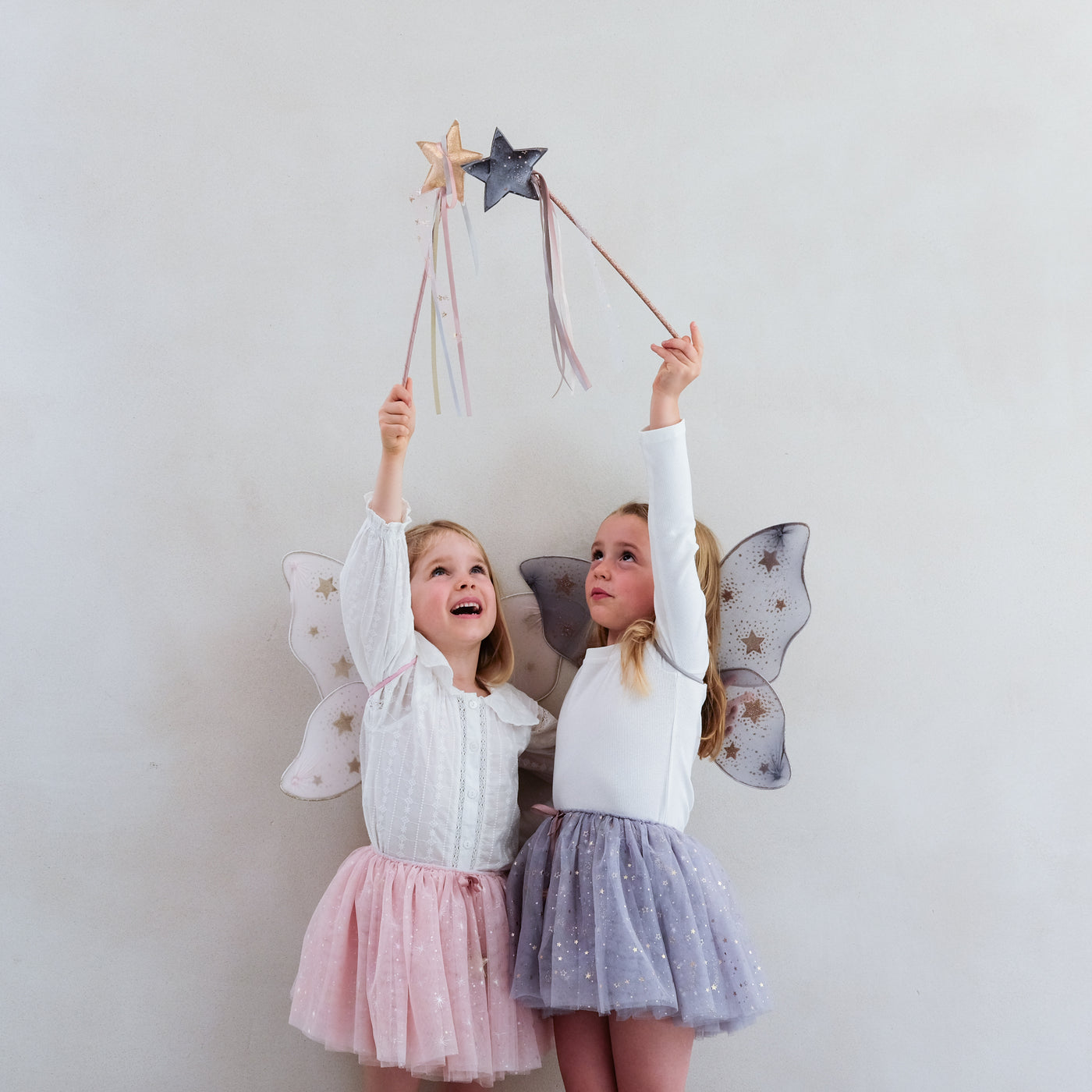 Little girl wearing grey tulle tutu speckled with gold stars and grey netted gold star print fairy wings and another wearing a pink tutu and pink wings, both with matching wands