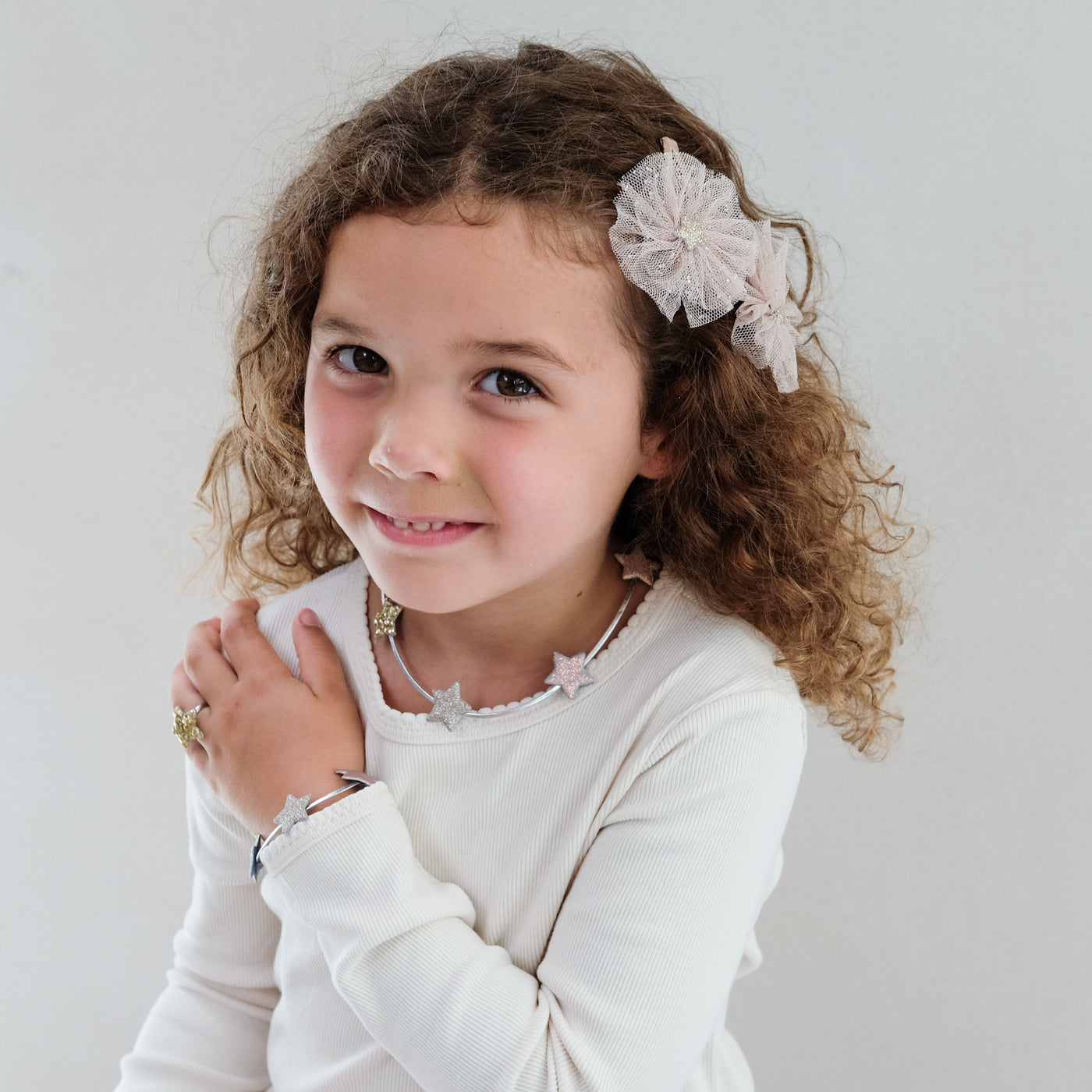 Young girl with curly hair wearing a star ring, star bracelet and star necklace, smiling against a plain background.