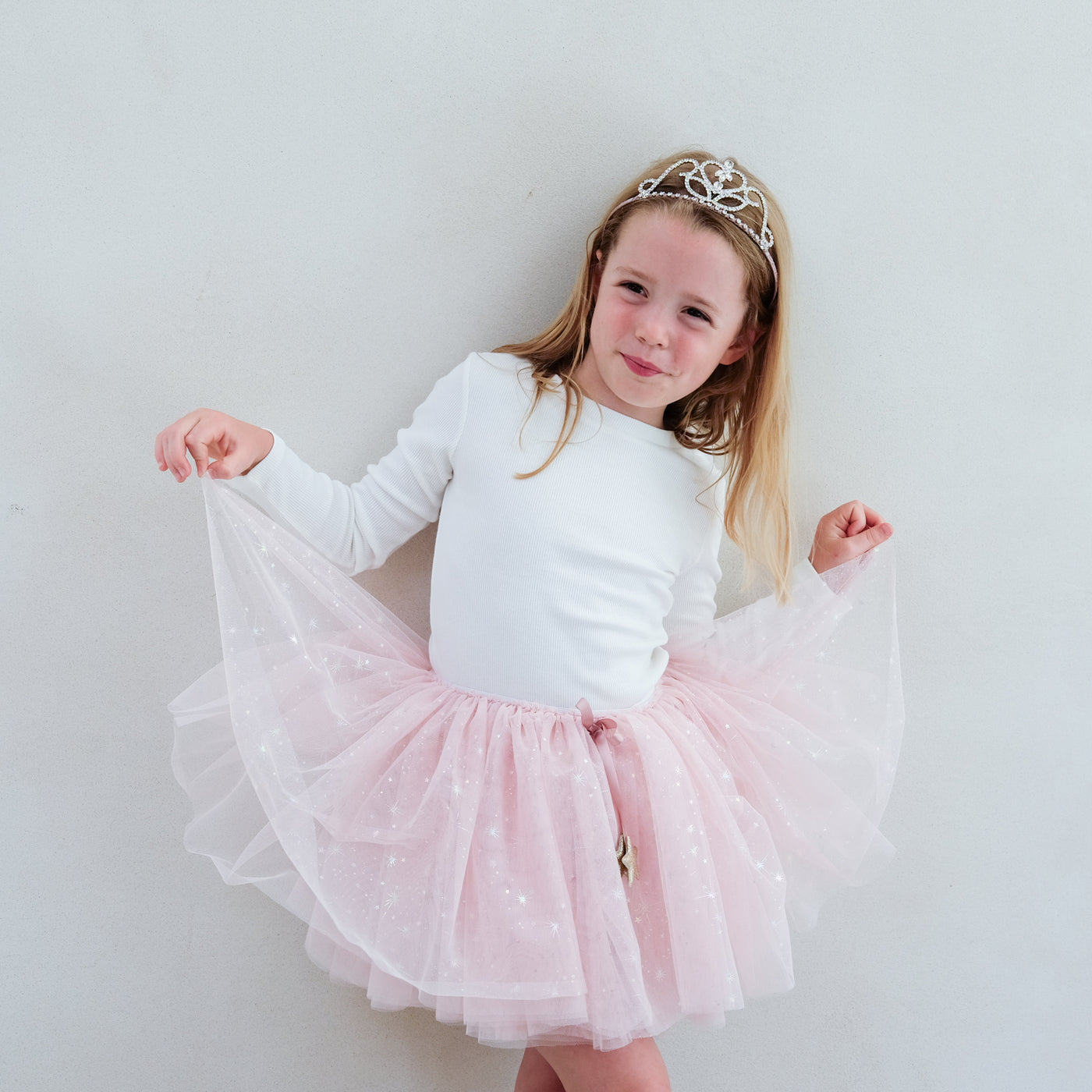 Young girl in a white long-sleeve top and pink tutu skirt with a tiara on a plain background