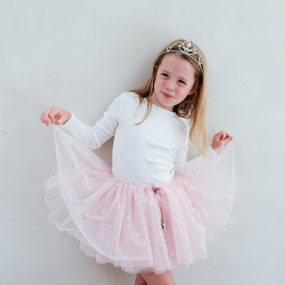 Young girl in a white long-sleeve top and pink tutu skirt with a tiara on a plain background