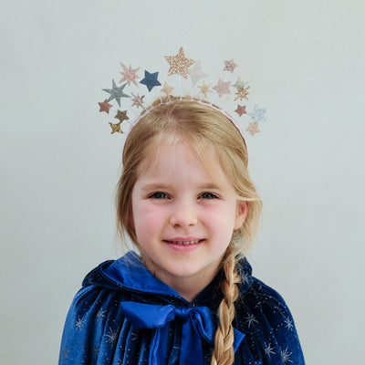 Young girl wearing a blue cape with star patterns and a star-shaped headband on a light grey background