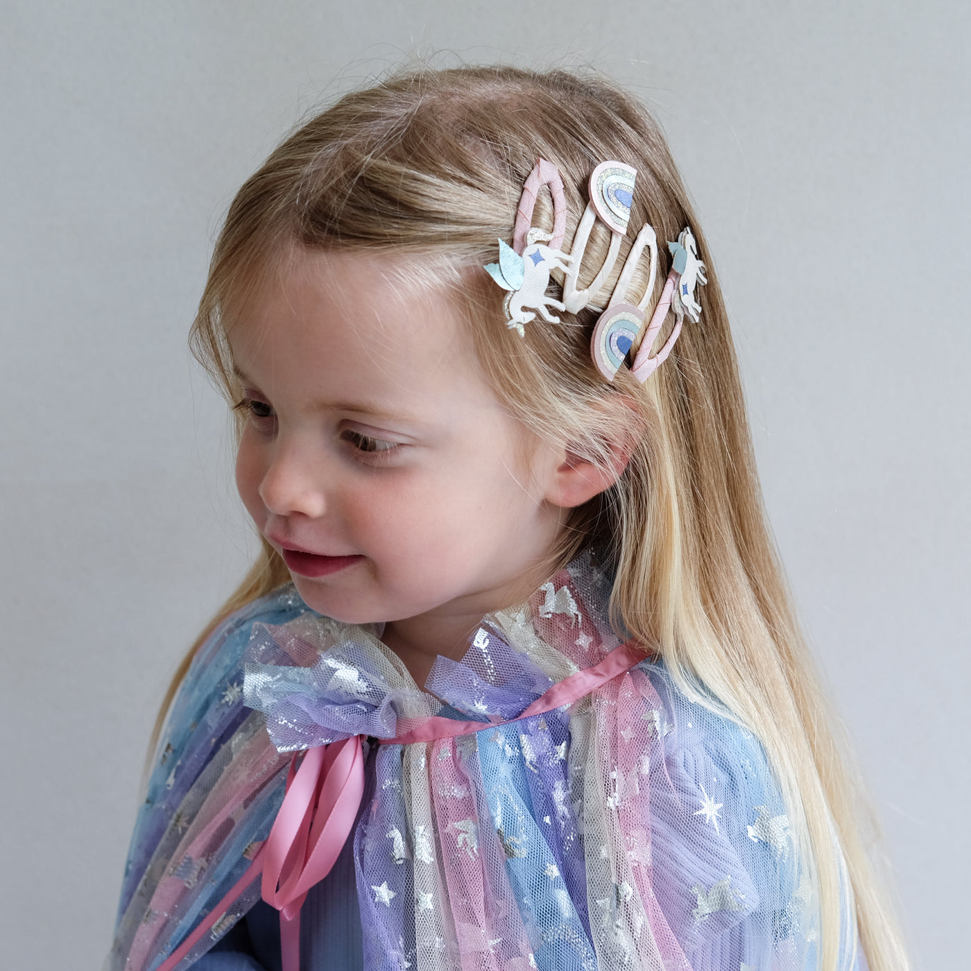 Young girl with unicorn hair clips and a colourful cape against a plain background