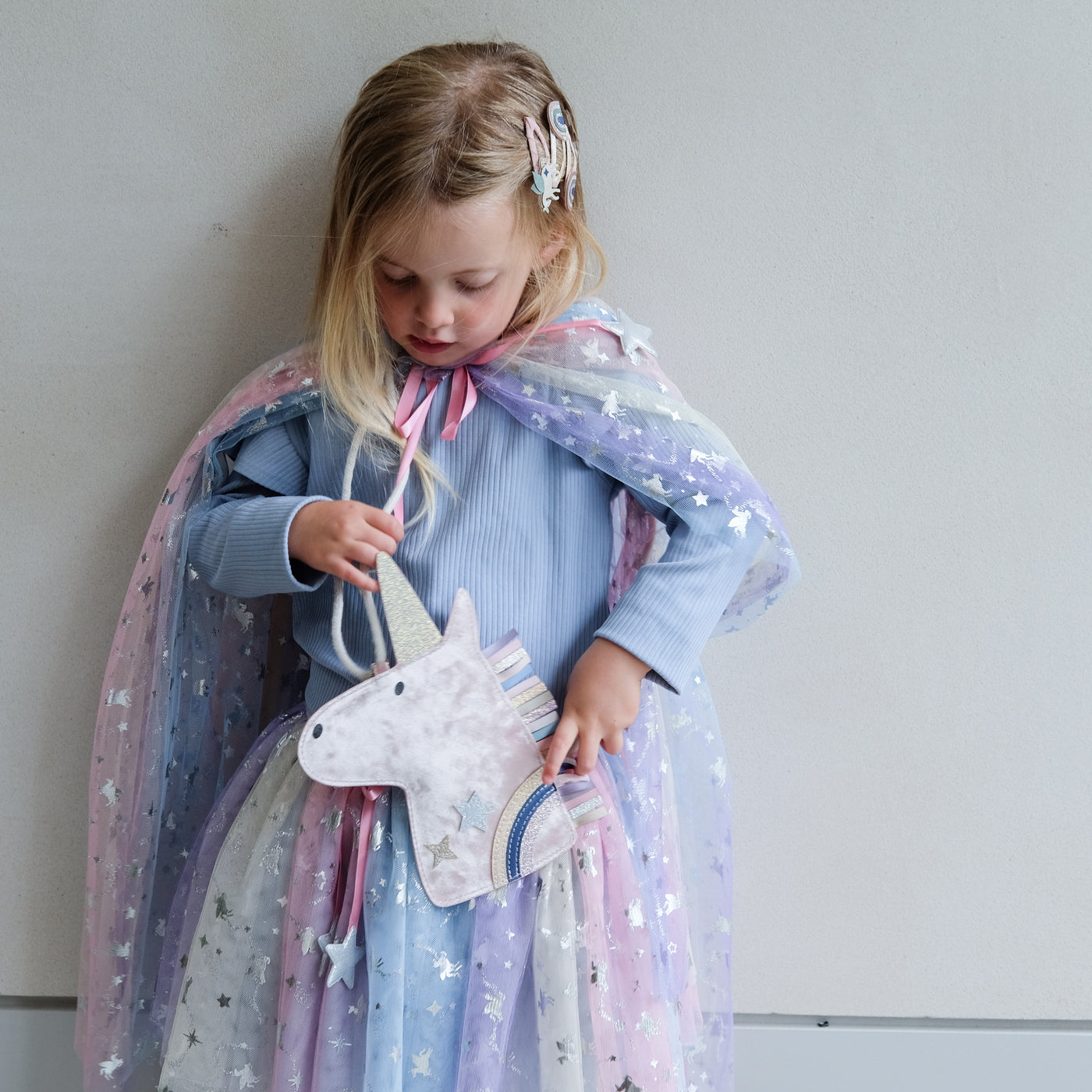Child wearing a star-patterned cape holding a unicorn bag against a plain background