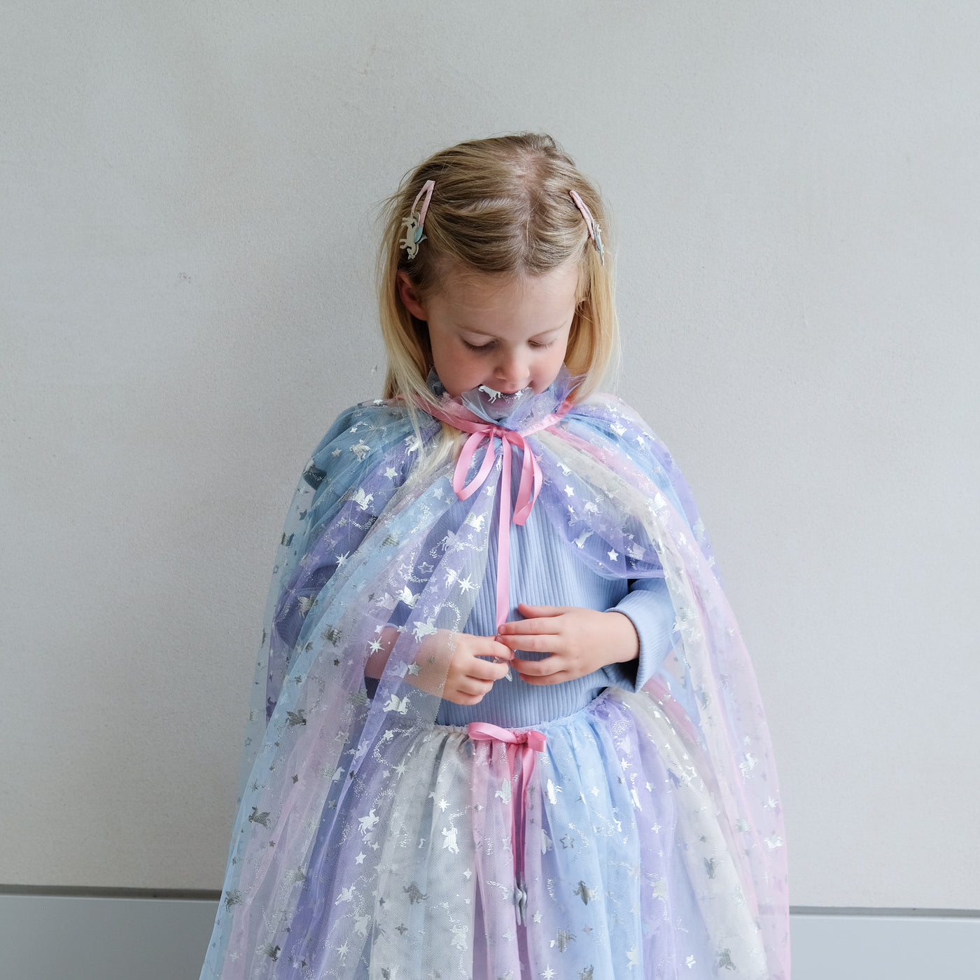 Young girl wearing a colourful star-patterned rainbow cape and tutu against a plain background