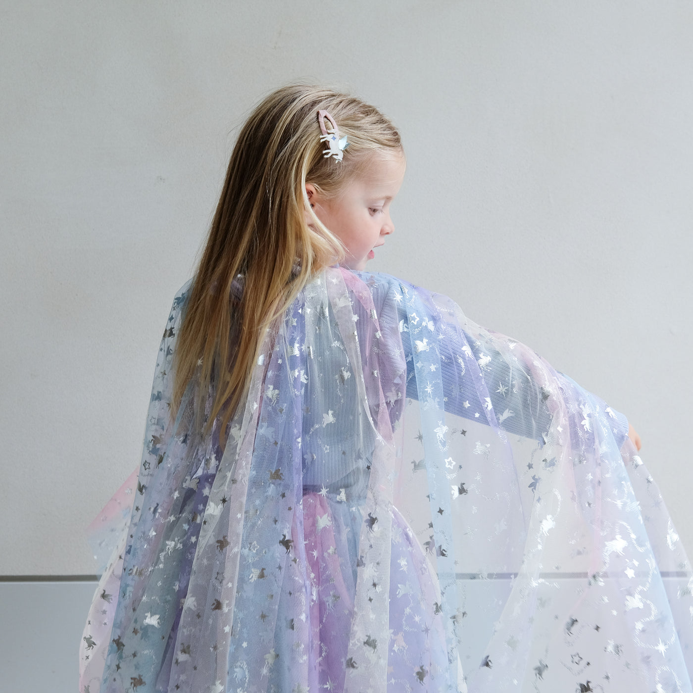 Young girl wearing a star-patterned rainbow cape against a plain background