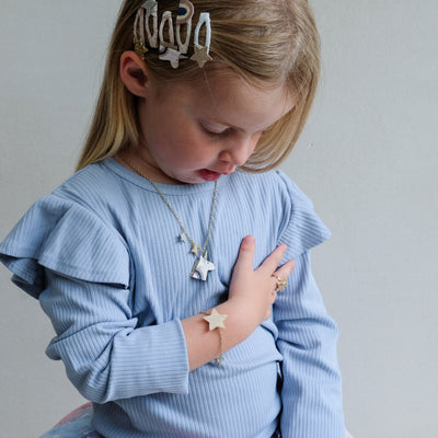 Young girl wearing a light blue sweater with ruffled sleeves, gold hair clip, necklace, and bracelet on a plain background.