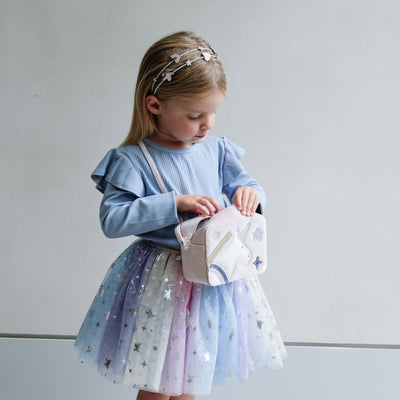 Young girl in a light blue sweater and colourful skirt holding a boxy bag with unicorn design against a white background