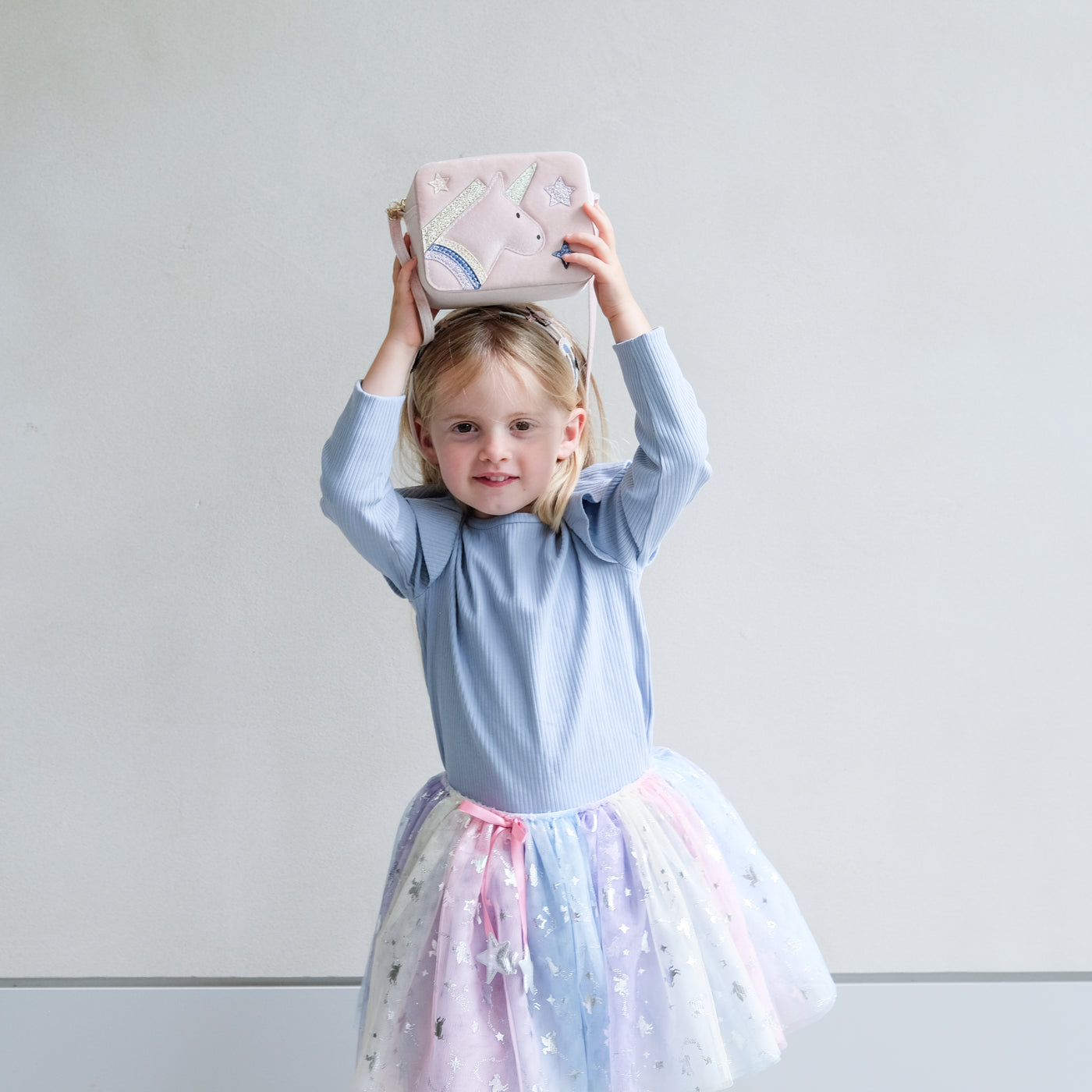 Young girl in a light blue top and colourful tutu holding a pink boxy bag with unicorn design against a plain background