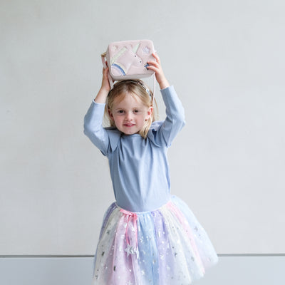 Young girl in a light blue top and colourful tutu holding a pink boxy bag with unicorn design against a plain background