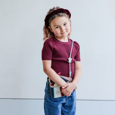 Young girl wearing a maroon sweater and blue jeans, holding a circus tent bag against a light background