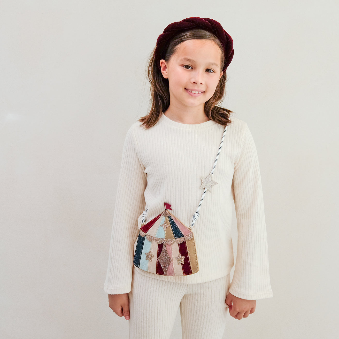 Young girl wearing a cream sweater and colourful circus tent handbag against a plain background