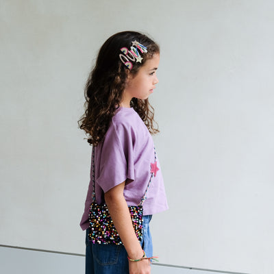Young girl wearing a purple shirt with colourful hair clips and sequin bag against a plain background