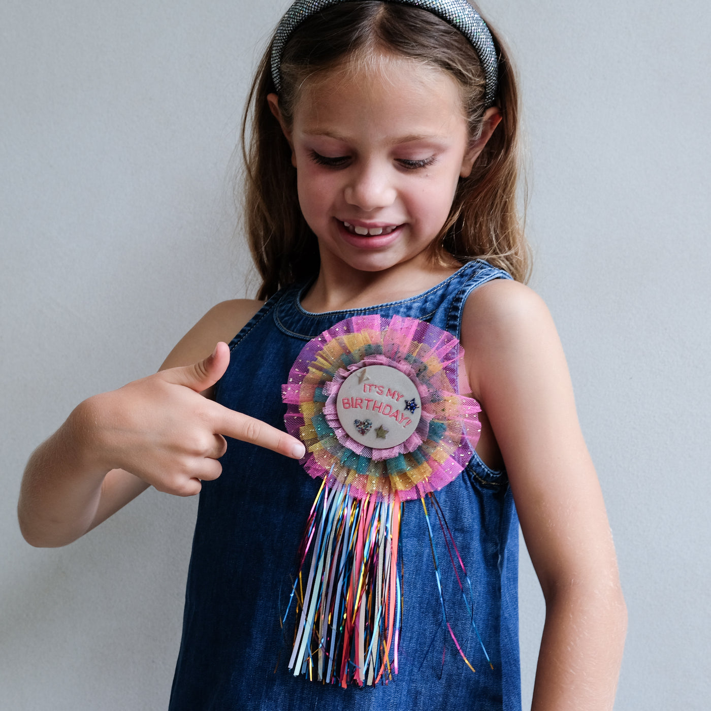 Young girl wearing a colourful birthday rosette badge on her top, pointing at it.