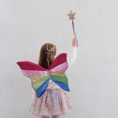 Child wearing colourful fairy wings and holding a star wand against a plain background
