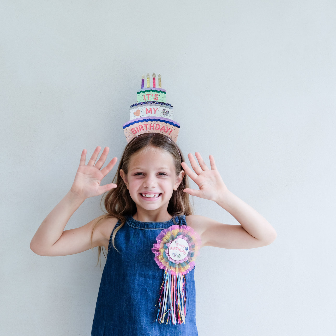Young girl wearing a colourful birthday headdress and holding up her hands against a light grey background