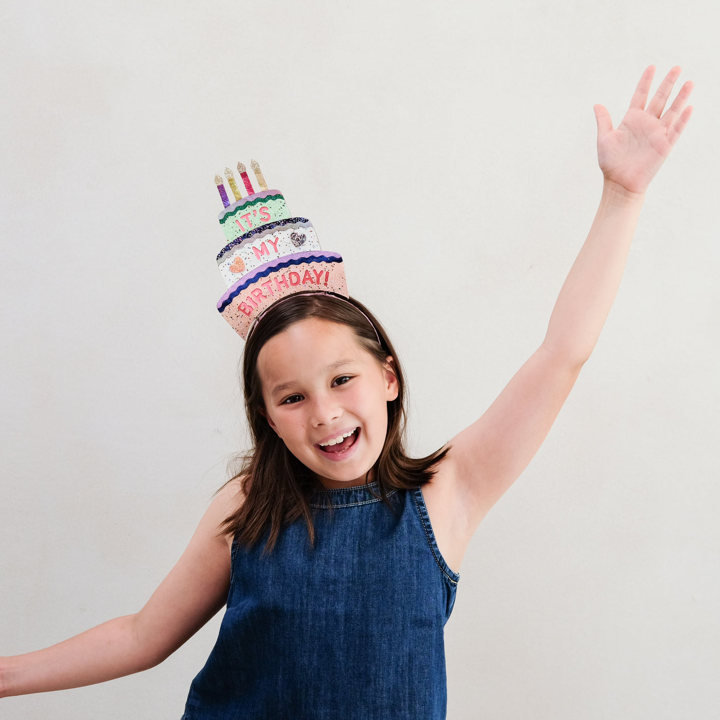 Child wearing a colourful birthday cake alice band with arms raised against a plain background