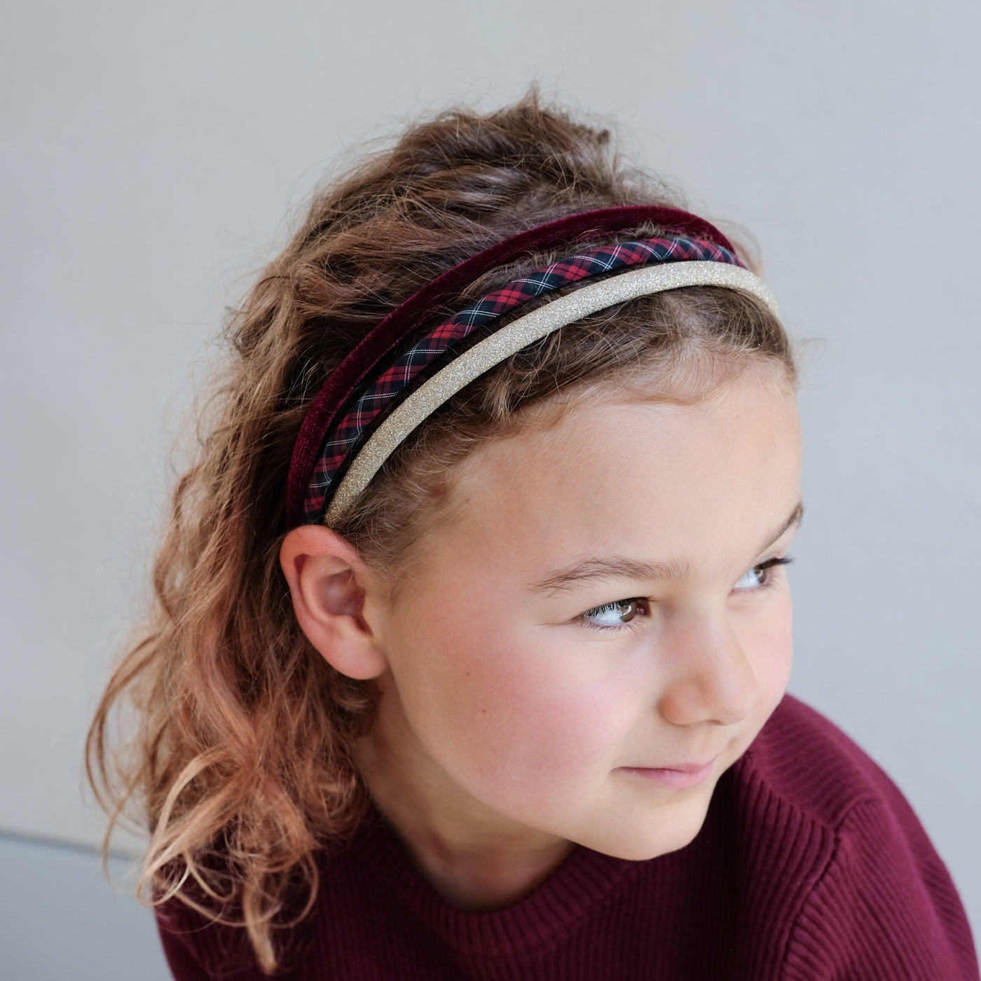 young girl wearing a pack of velvet, tartan, and gold glitter headband against a white background