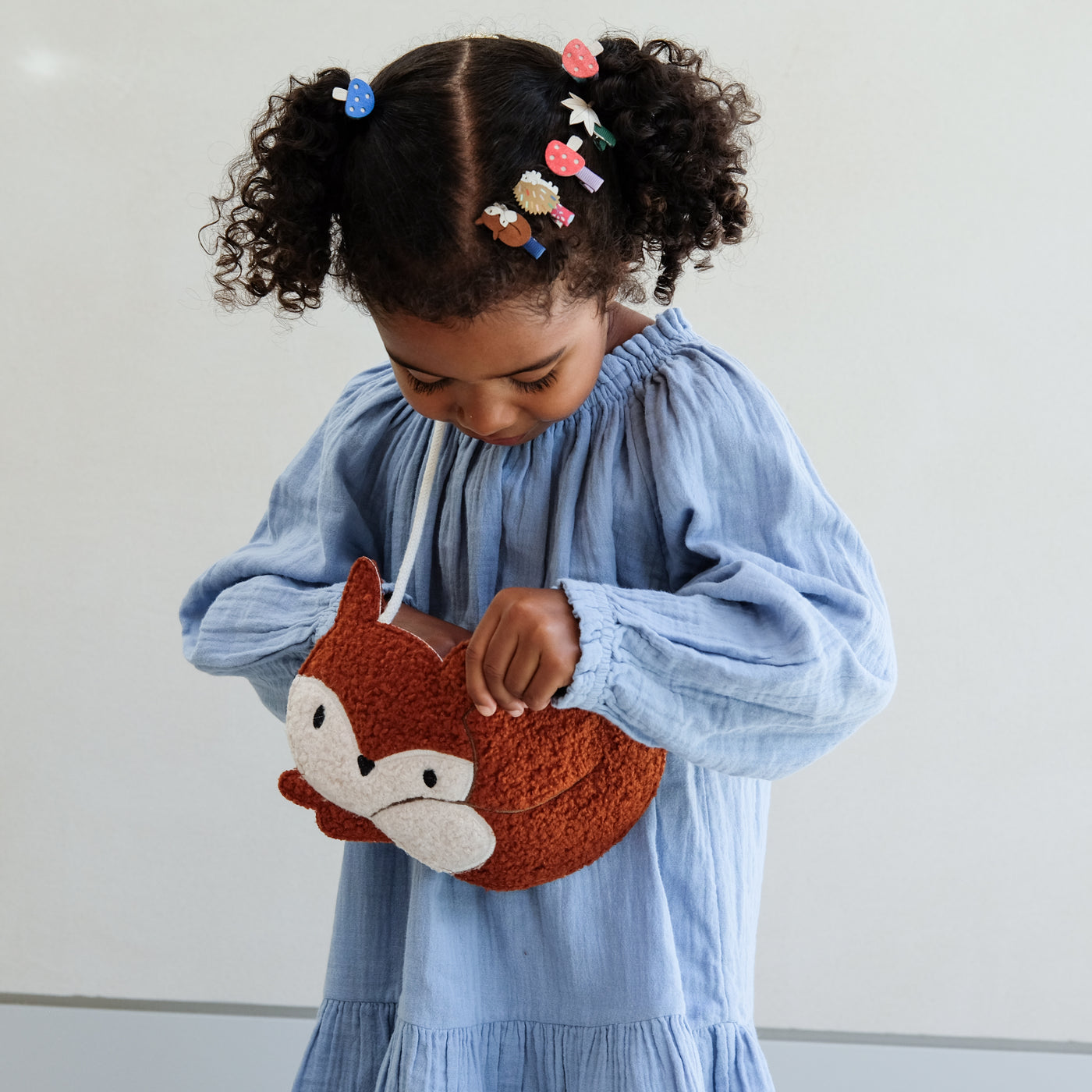 Child holding a fox-shaped bag against a plain background with girls hair clips and hair ties in her hair