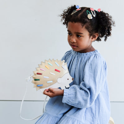 Child holding a colorful hedgehog-shaped bag with girls hair clips in her hair against a light background