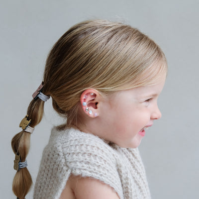 Young girl with braided hair and decorative gem stick on earrings wearing a light-coloured knit garment against a neutral background