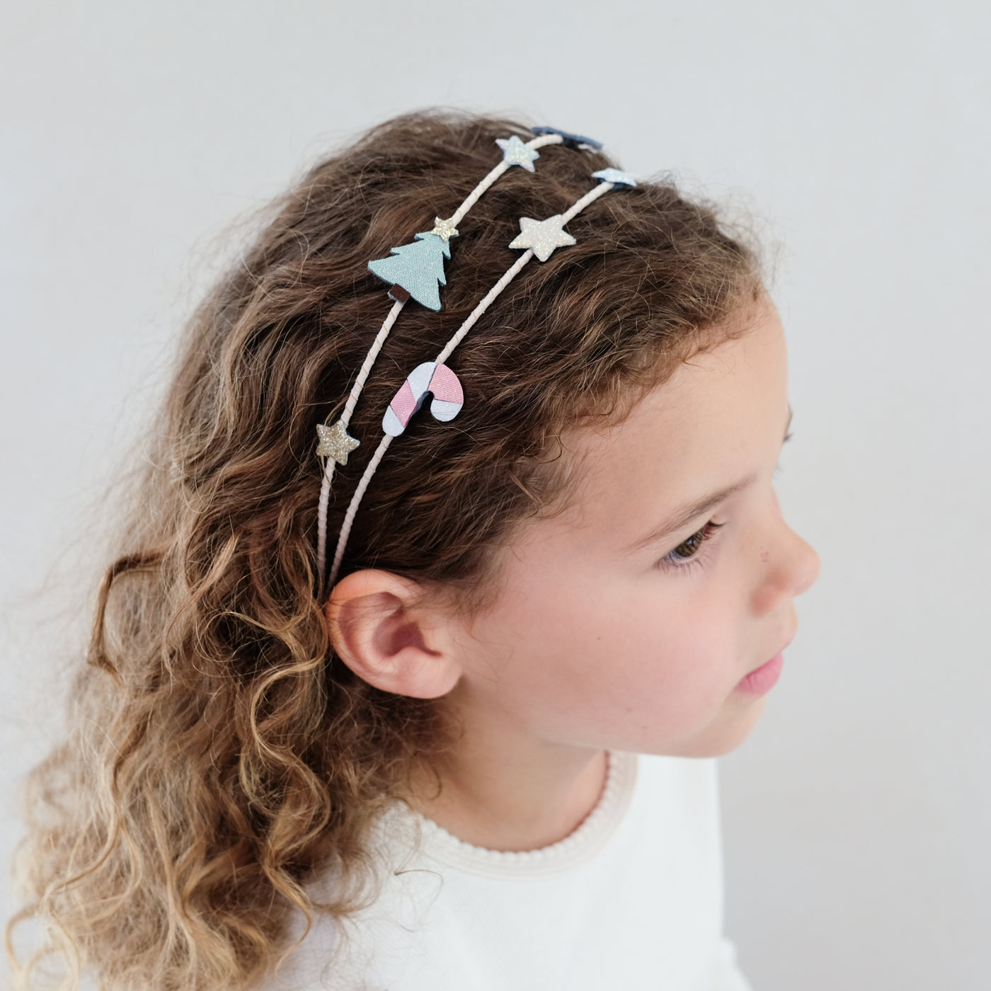 Young girl wearing a decorative headband with candy cane and a Christmas tree on a plain background
