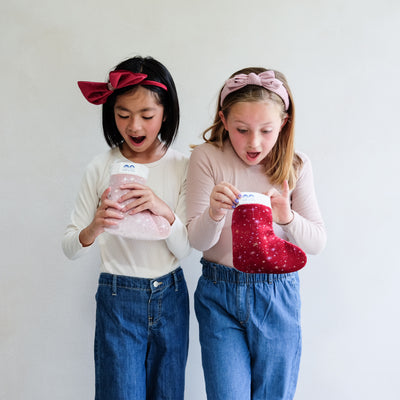 two young girls holding a pink christmas gift stocking and a red christmas gift stocking against a plain white background