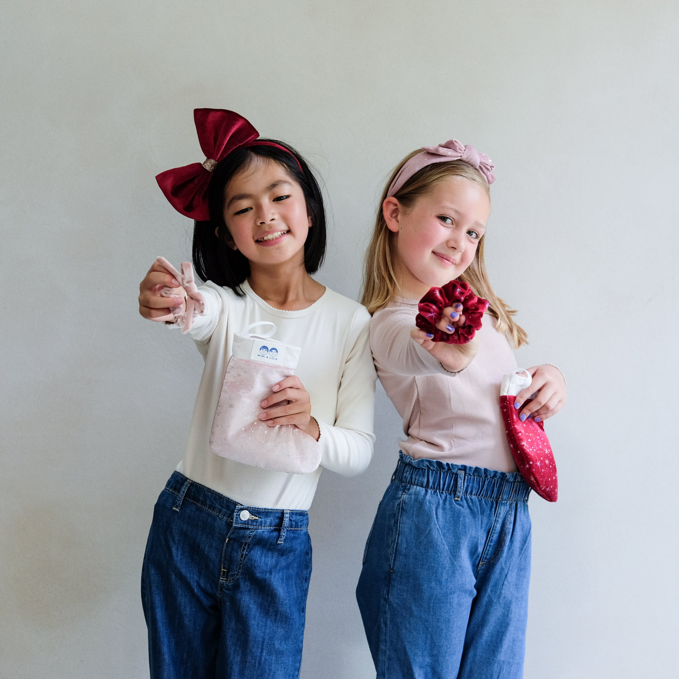 two young girls holding a pink christmas gift stocking and a red christmas gift stocking against a plain white background