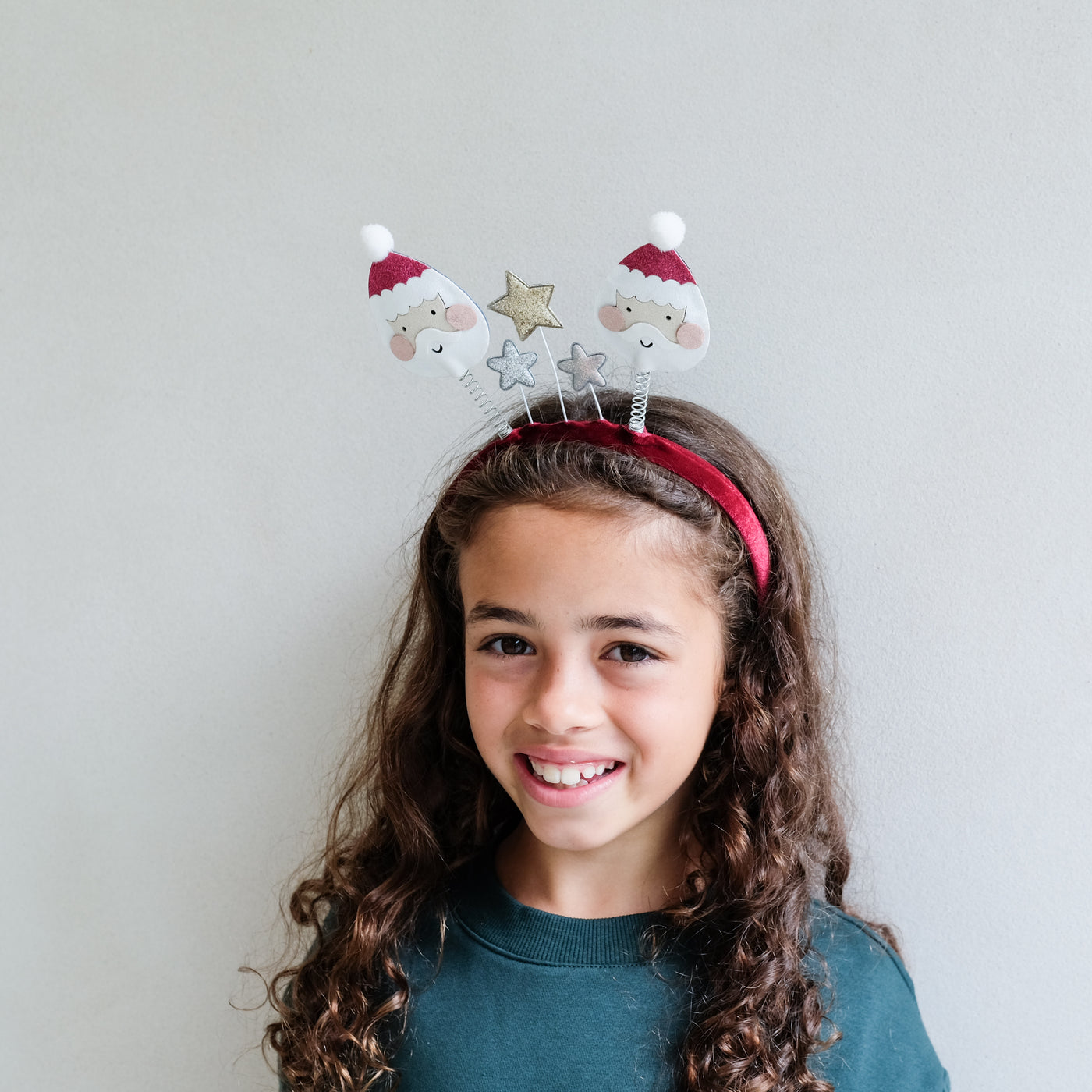 Young girl wearing a festive headband with Santa faces and stars on a plain background