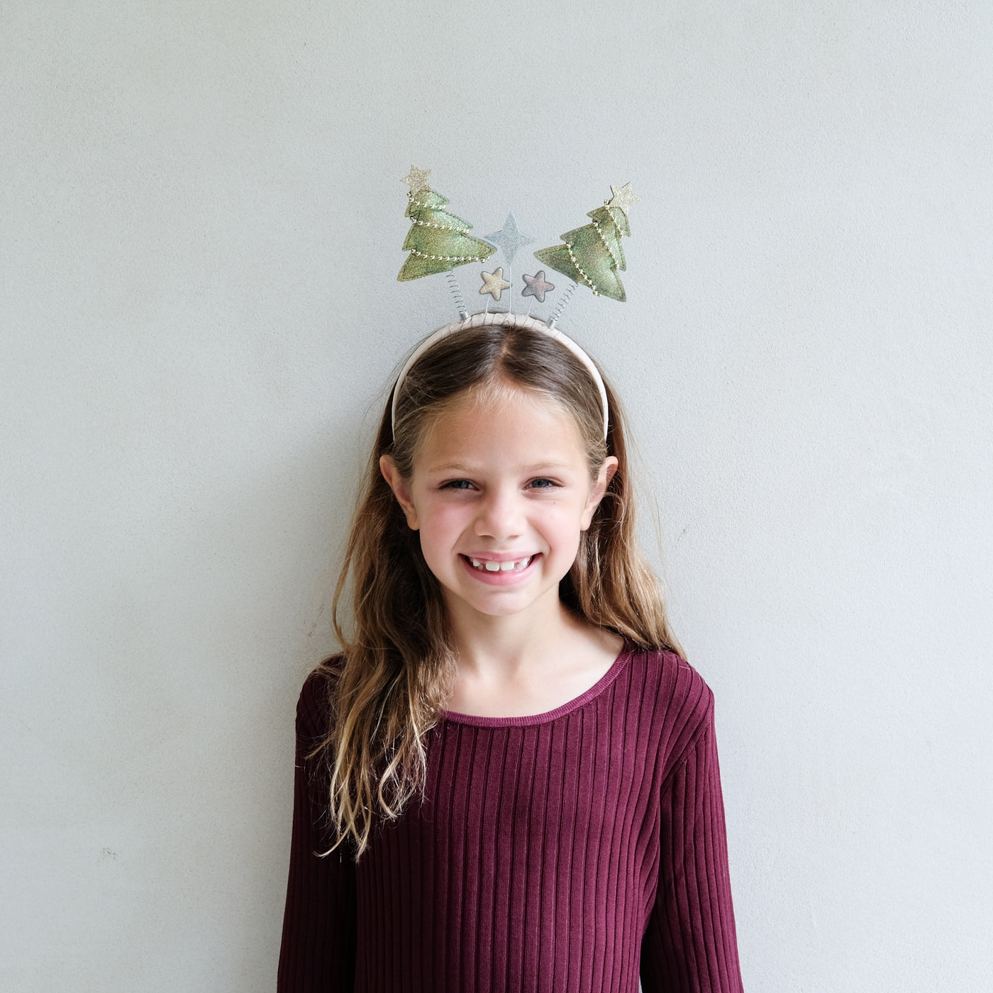 young girl wearing a Christmas tree bopper headband against a white background