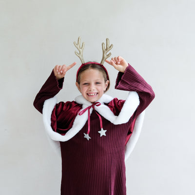 Child wearing a Santa velvet red cape and gold glitter reindeer antlers against a plain background