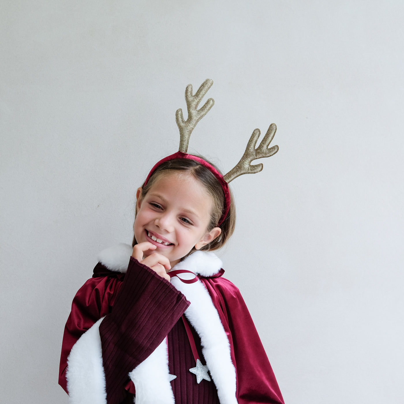 Child wearing a Santa velvet red cape and gold glitter reindeer antlers against a plain background
