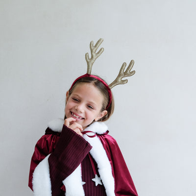 Child wearing a Santa velvet red cape and gold glitter reindeer antlers against a plain background