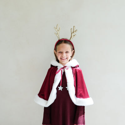 Child wearing a red dress with white fur trim, a red cape, and gold antlers against a plain background
