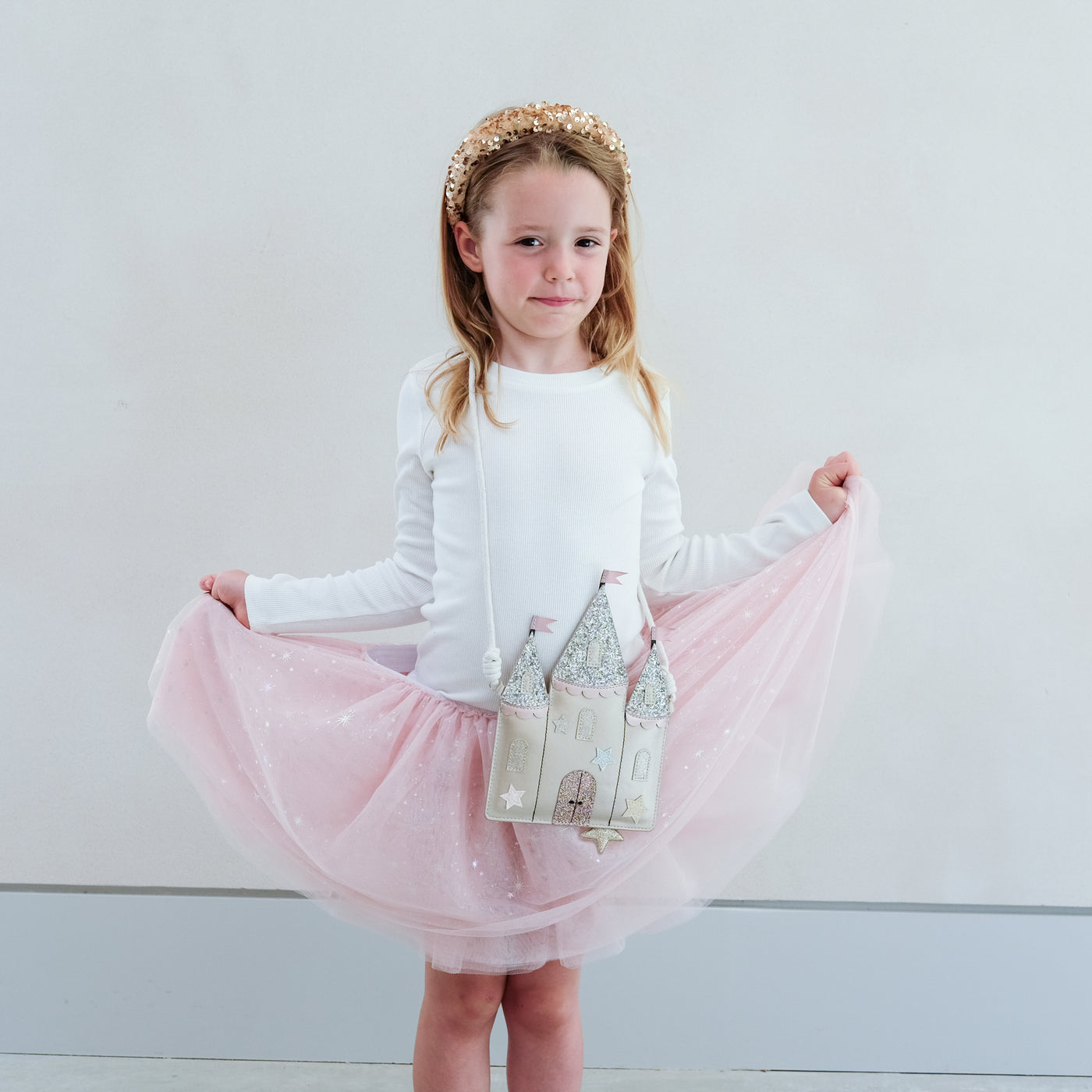 Young girl in a white top and pink skirt holding a castle-shaped bag against a plain background