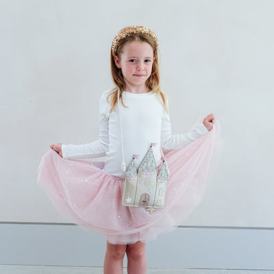 Young girl in a white top and pink skirt holding a castle-shaped bag against a plain background
