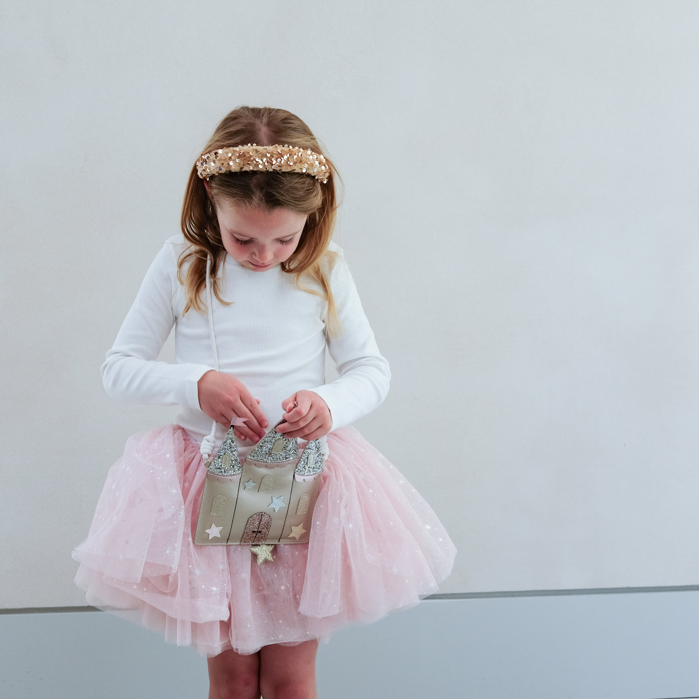 Young girl in a white long-sleeve top and pink tutu skirt holding a castle shaped bag against a plain background.