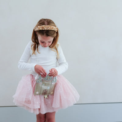 Young girl in a white long-sleeve top and pink tutu skirt holding a castle shaped bag against a plain background.