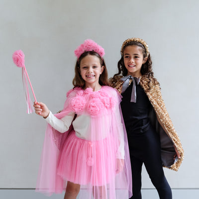 Young girl wearing a pink ruffle floral dressing up outfit and floral headband against a white background, and another young girl wearing a gold sequin cape and sequin headband