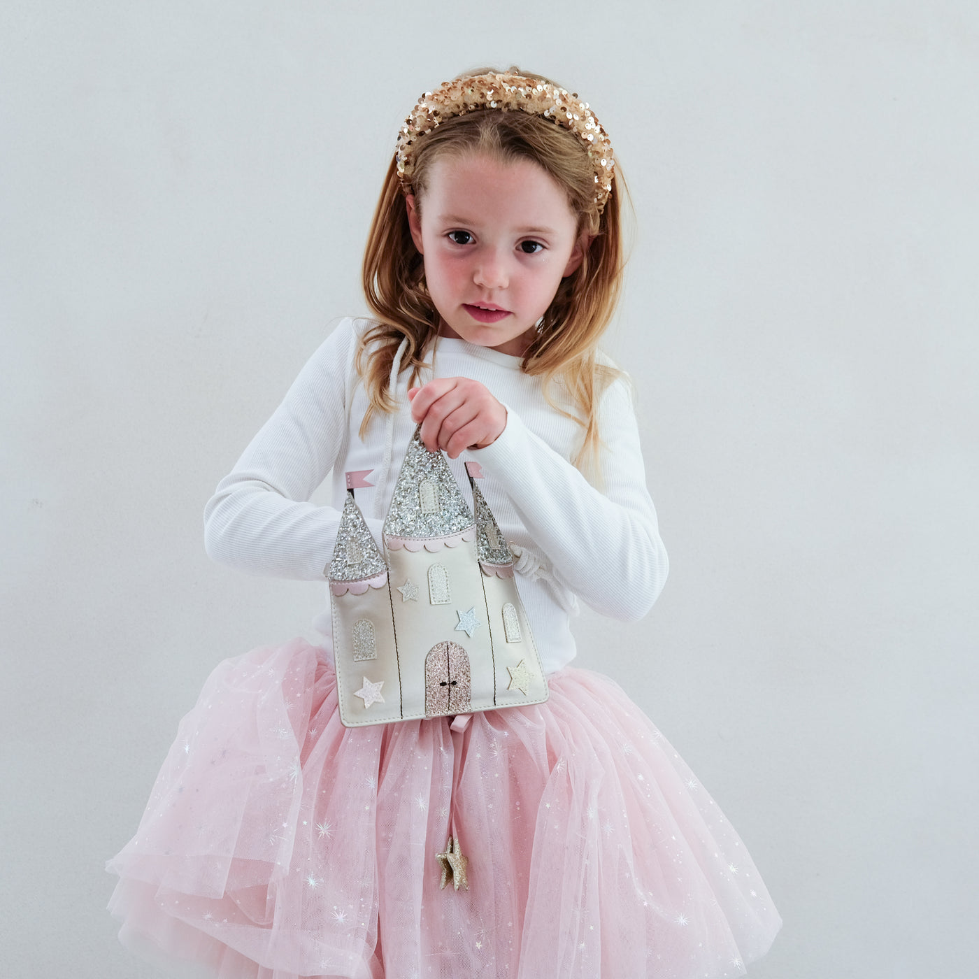 Young girl in a pink tutu dress with a white long-sleeve top and a decorative castle shaped bag, standing against a plain background.