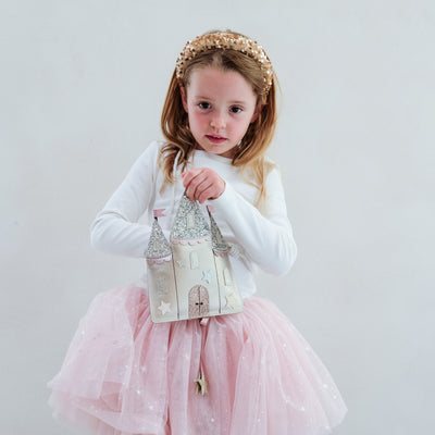 Young girl in a pink tutu dress with a white long-sleeve top and a decorative castle shaped bag, standing against a plain background.